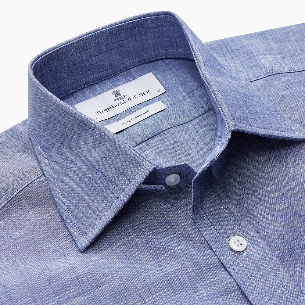 The Turnbull & Asser Blue Chambray Cotton Regular Fit Mayfair Shirt features a subtle textured pattern, pointed T&A collar, and white buttons. Shown here in a light blue shade, laid flat on a white background.