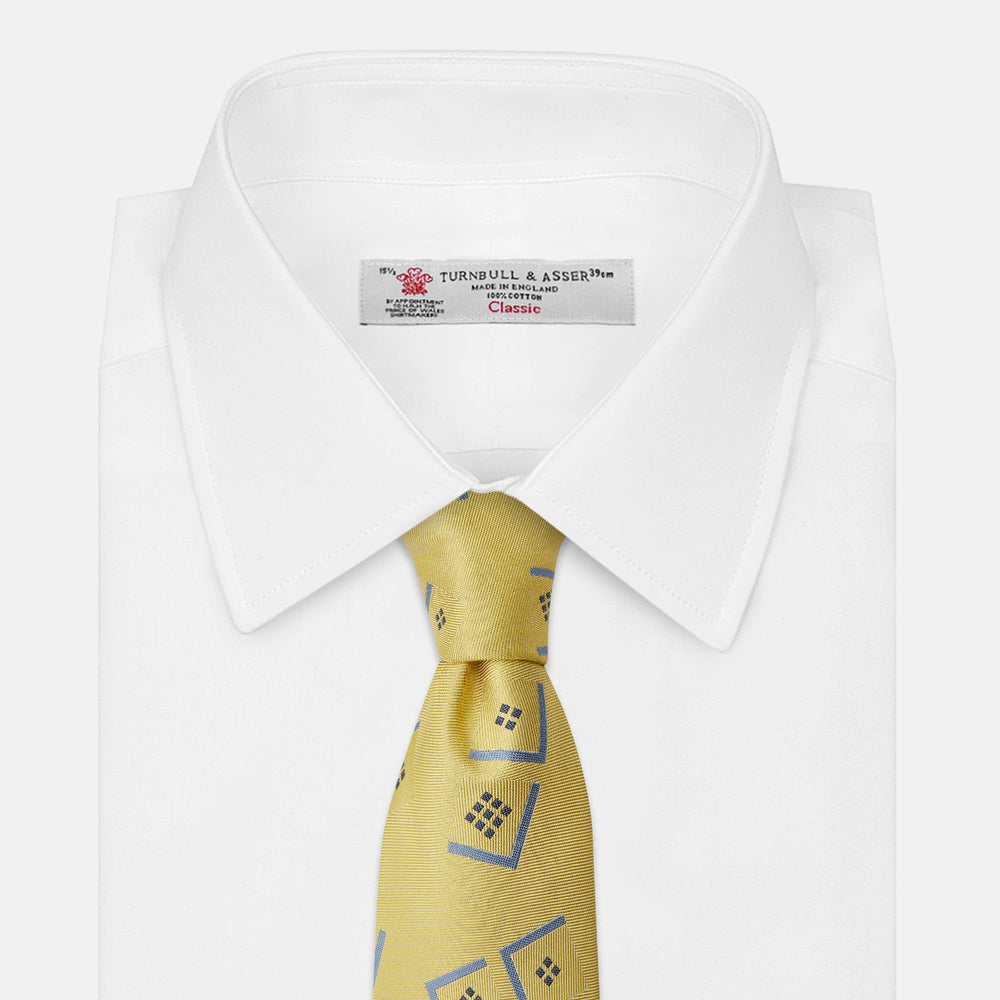 The Turnbull & Asser Yellow Graphic Deco Silk Tie features a subtle pattern of light blue geometric shapes and small black dots, elegantly rolled against a white background.