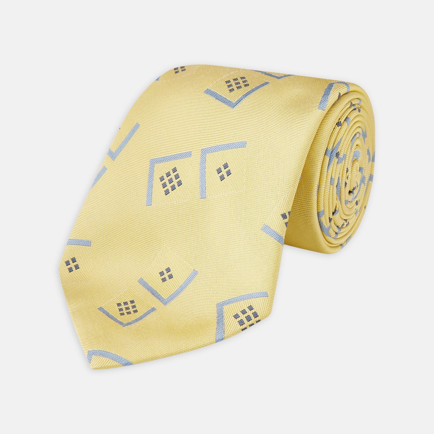 View 1 of product The Turnbull & Asser Yellow Graphic Deco Silk Tie features a subtle pattern of light blue geometric shapes and small black dots, elegantly rolled against a white background.