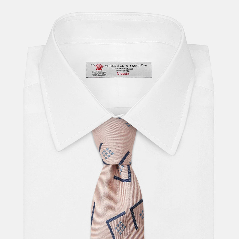 The Turnbull & Asser Pink Graphic Deco Silk Tie features blue geometric and square patterns on pale pink silk, shown rolled with the pointed end facing forward.