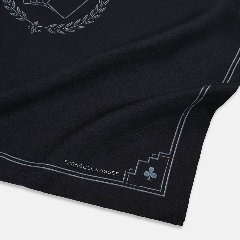 Introducing the Casino Royale Midnight Blue Crest Pocket Square in Silk from Turnbull & Asser UK—a midnight blue silk pocket square bordered in white, featuring playing card suit symbols at each corner and a central motif depicting the 10, J, Q, and K of spades beneath a crown and laurel. Perfect as a sophisticated Casino Royale or Bond tuxedo accessory, with “Turnbull & Asser” elegantly displayed in the lower right.
