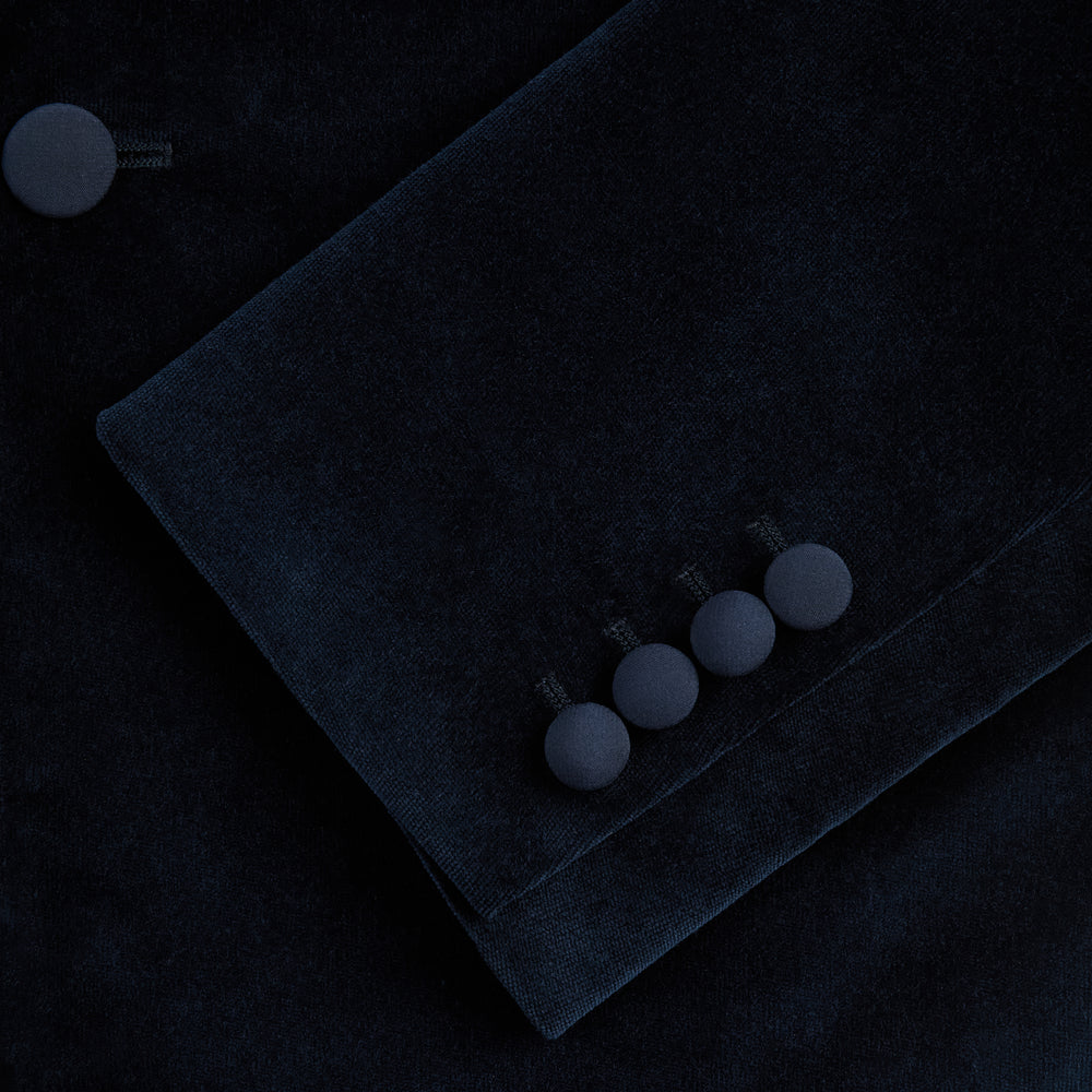 Turnbull & Asser UK’s Casino Royale Midnight Blue Tuxedo Jacket in Cotton Velvet, featuring a single button closure and a luxurious silk-satin shawl lapel, is showcased against a plain white background.