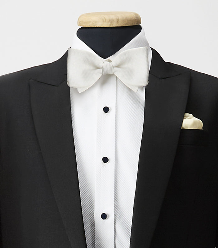 A black tuxedo jacket with satin lapels, a white dress shirt with black studs, a white bow tie, and a cream pocket square displayed on a mannequin.