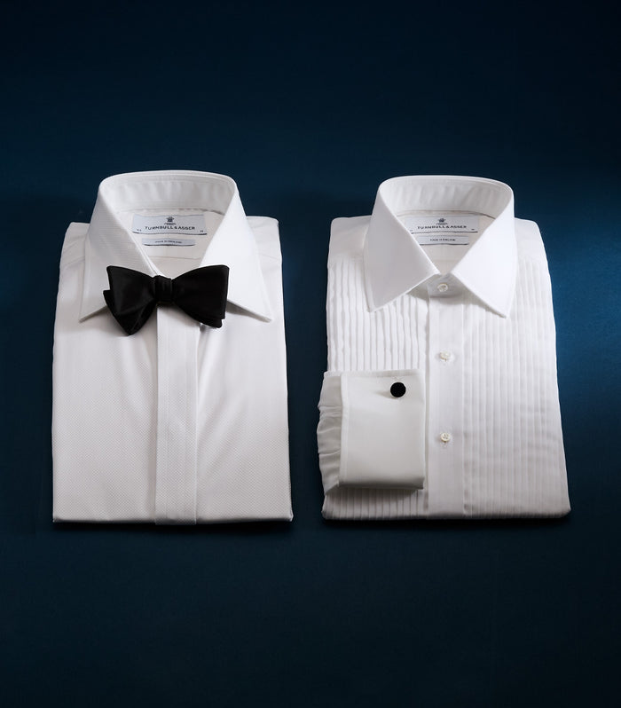 Two neatly folded white dress shirts are side by side on a dark blue surface; the left shirt has a black bow tie, while the right features pleated fabric and a French cuff with a black cufflink.