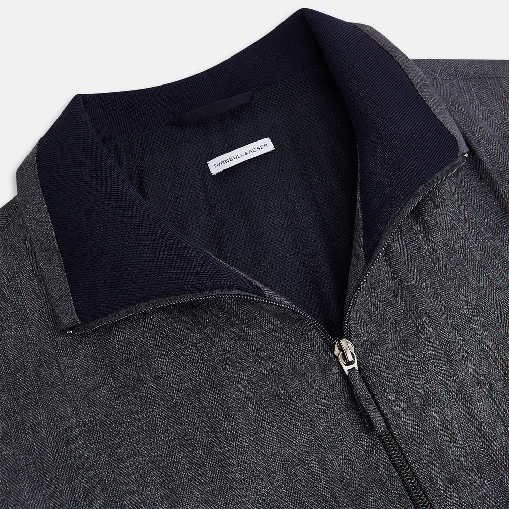 A front view of the Fairway Ash Golf Jacket in Linen by Turnbull & Asser UK is shown, featuring a high collar, two side zip pockets, a chest zip pocket, and black ribbed cuffs and hem. Crafted from lightweight gray herringbone linen for effortless warm-weather dressing, the jacket is displayed on a plain white background.