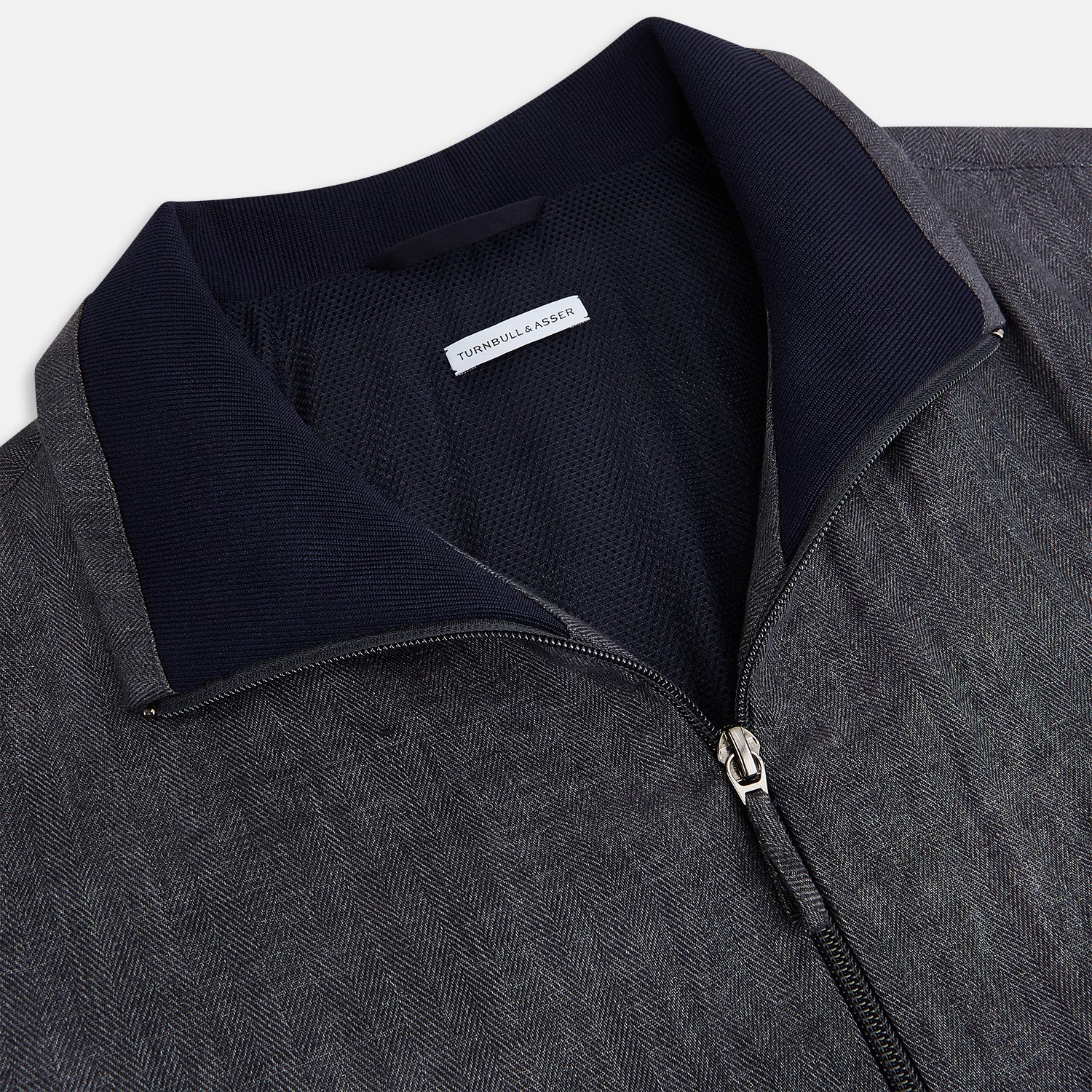 View 2 of product A front view of the Fairway Ash Golf Jacket in Linen by Turnbull & Asser UK is shown, featuring a high collar, two side zip pockets, a chest zip pocket, and black ribbed cuffs and hem. Crafted from lightweight gray herringbone linen for effortless warm-weather dressing, the jacket is displayed on a plain white background.