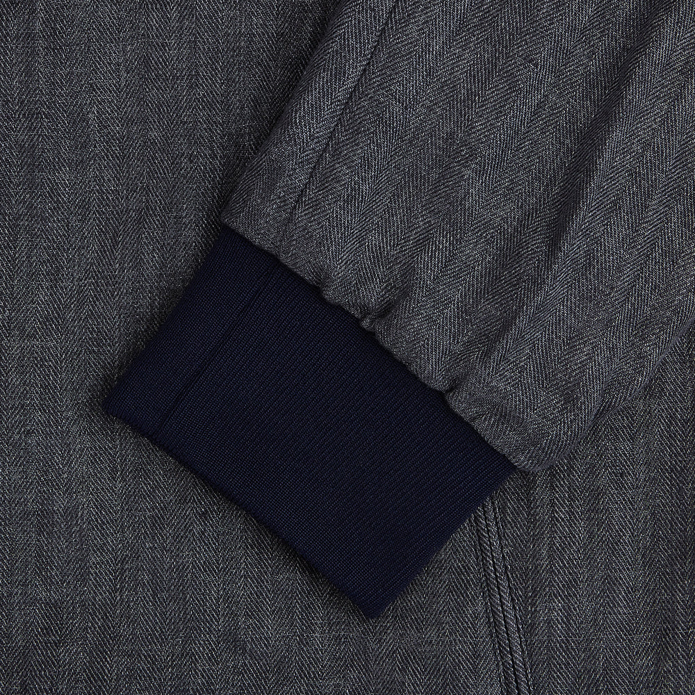 A front view of the Fairway Ash Golf Jacket in Linen by Turnbull & Asser UK is shown, featuring a high collar, two side zip pockets, a chest zip pocket, and black ribbed cuffs and hem. Crafted from lightweight gray herringbone linen for effortless warm-weather dressing, the jacket is displayed on a plain white background.