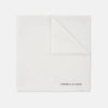 View 1 of product The White Piped Silk Pocket Square by Turnbull & Asser is shown folded at one corner with the brand's logo in black on the lower right, set against a light blue background.