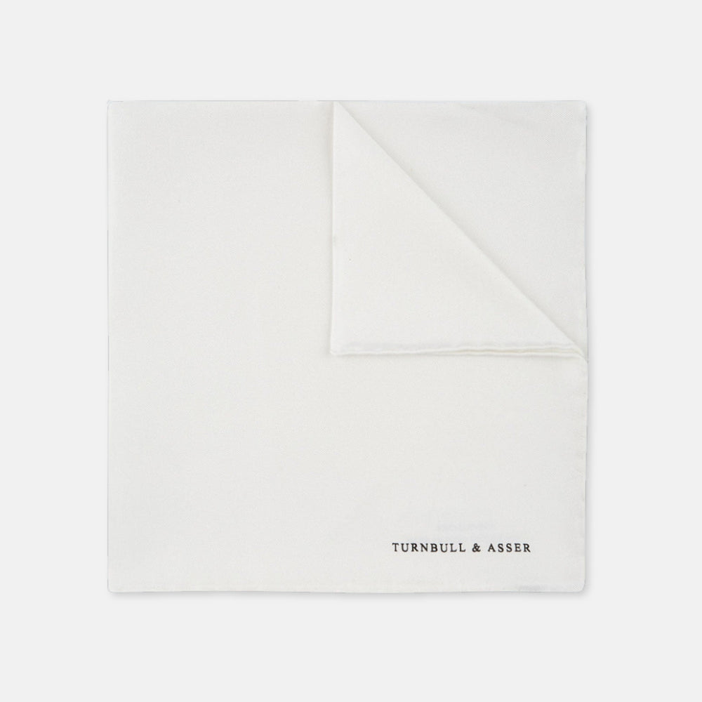 The White Piped Silk Pocket Square by Turnbull & Asser is shown folded at one corner with the brand's logo in black on the lower right, set against a light blue background.