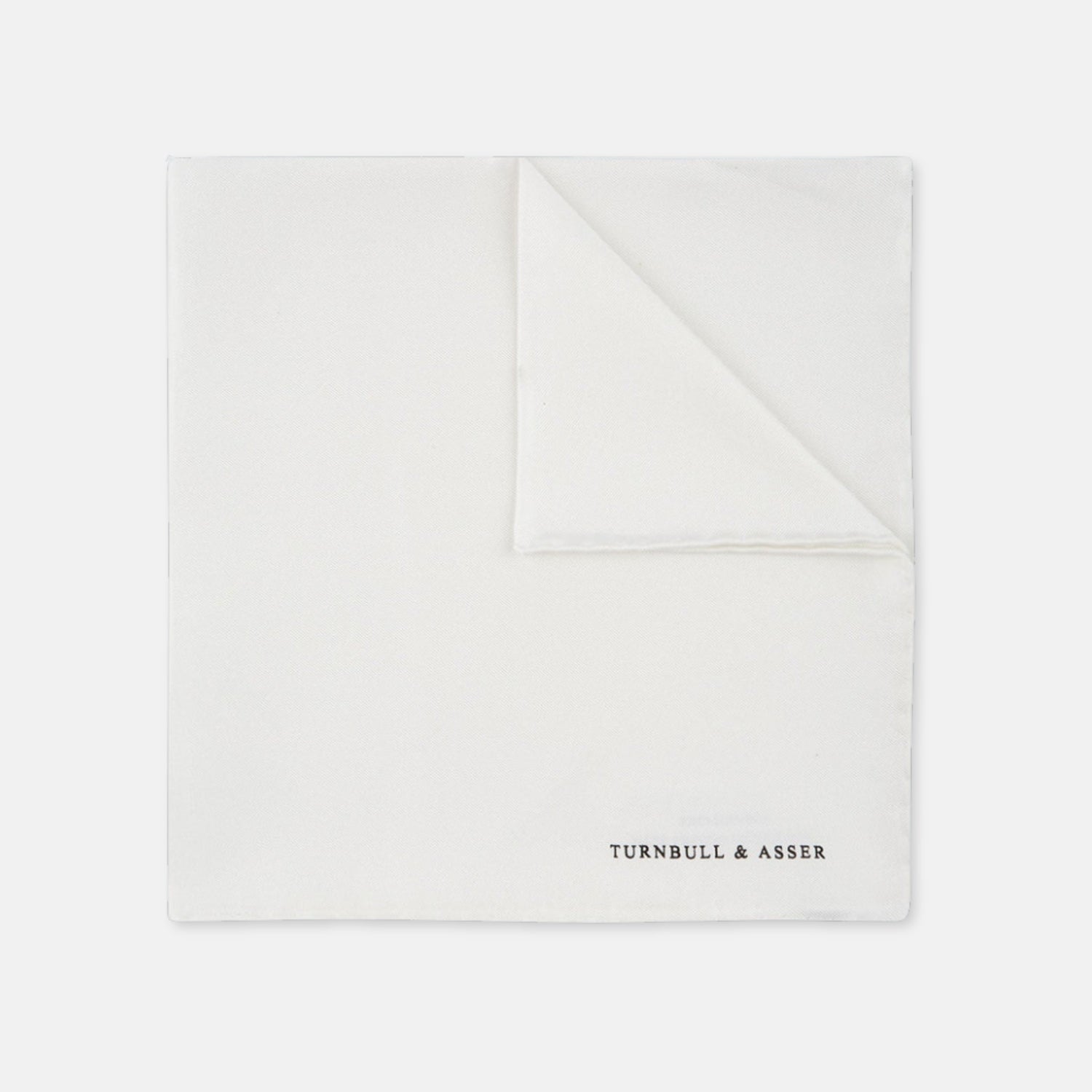 View 3 of product The White Piped Silk Pocket Square by Turnbull & Asser is shown folded at one corner with the brand's logo in black on the lower right, set against a light blue background.