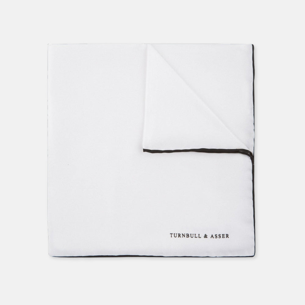 The Turnbull & Asser White and Black Piped Silk Pocket Square features a crisp white base with black edging, neatly folded to showcase the logo in black on the lower right—elegant monochrome style for any occasion, shown on a light gray background.