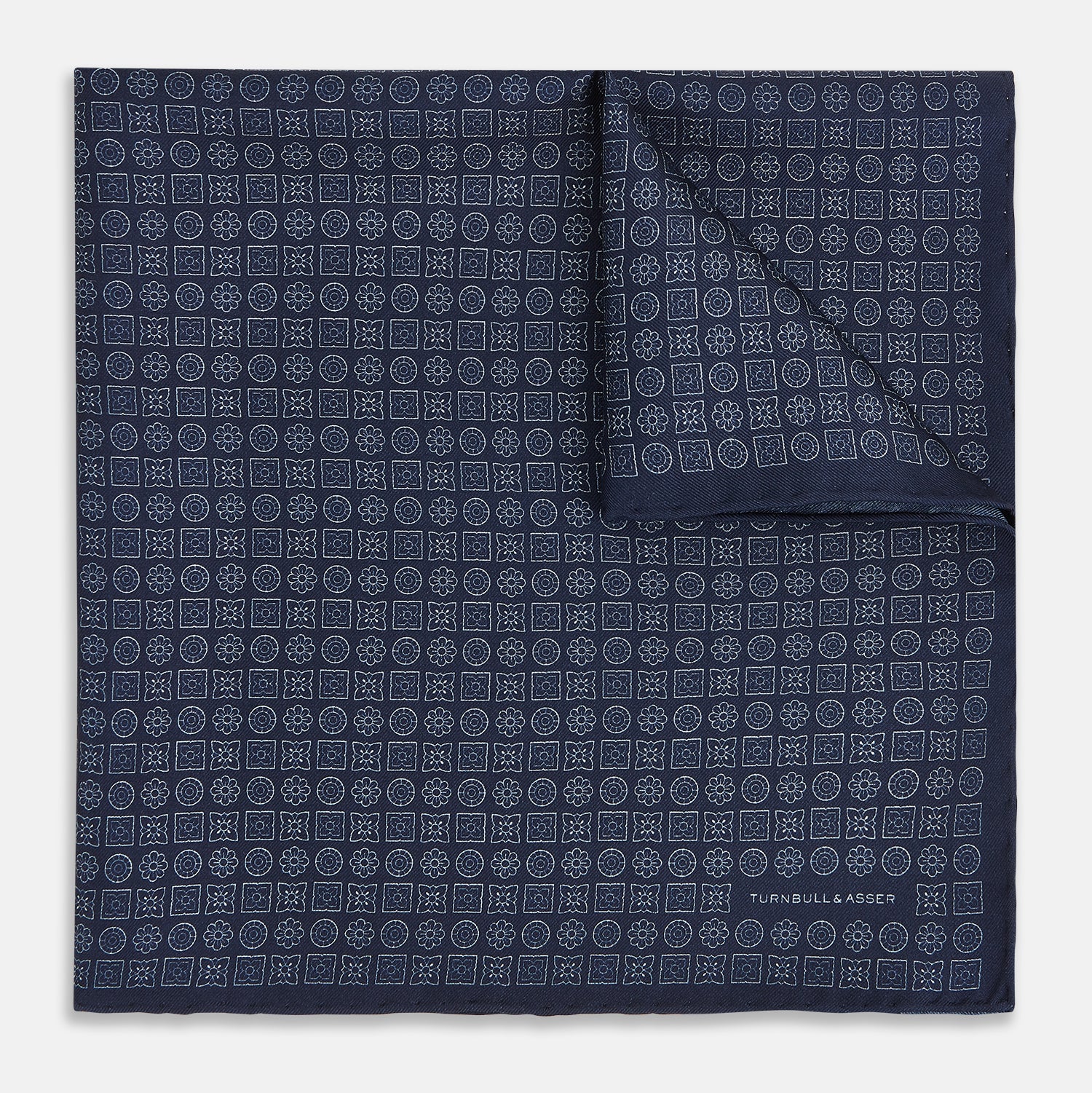 View 1 of product A Floral Navy Pocket Square in Silk by Turnbull & Asser UK, featuring a repeating geometric pattern with the brand’s name printed in white in the corner—perfect for adding a touch of sophistication to formal tailoring. The top right edge is folded over to reveal the reverse side.
