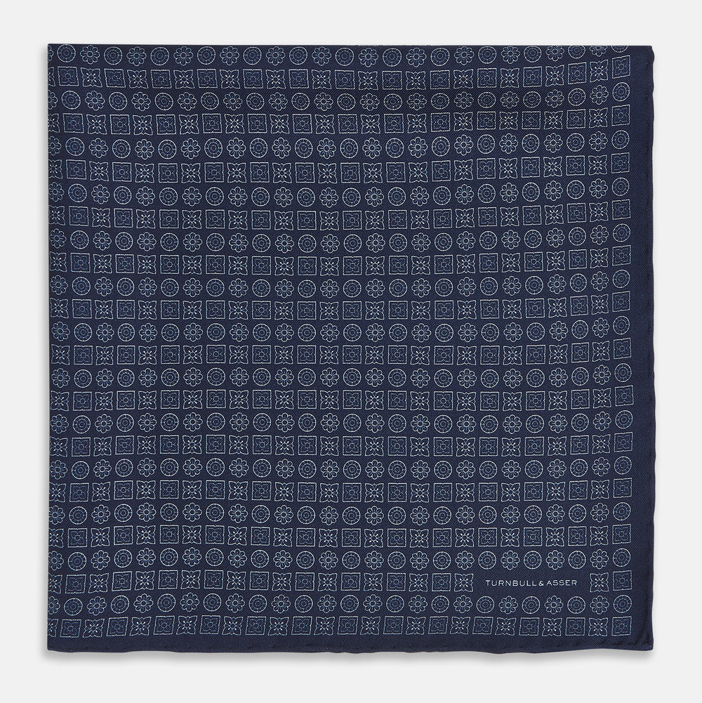A Floral Navy Pocket Square in Silk by Turnbull & Asser UK, featuring a repeating geometric pattern with the brand’s name printed in white in the corner—perfect for adding a touch of sophistication to formal tailoring. The top right edge is folded over to reveal the reverse side.