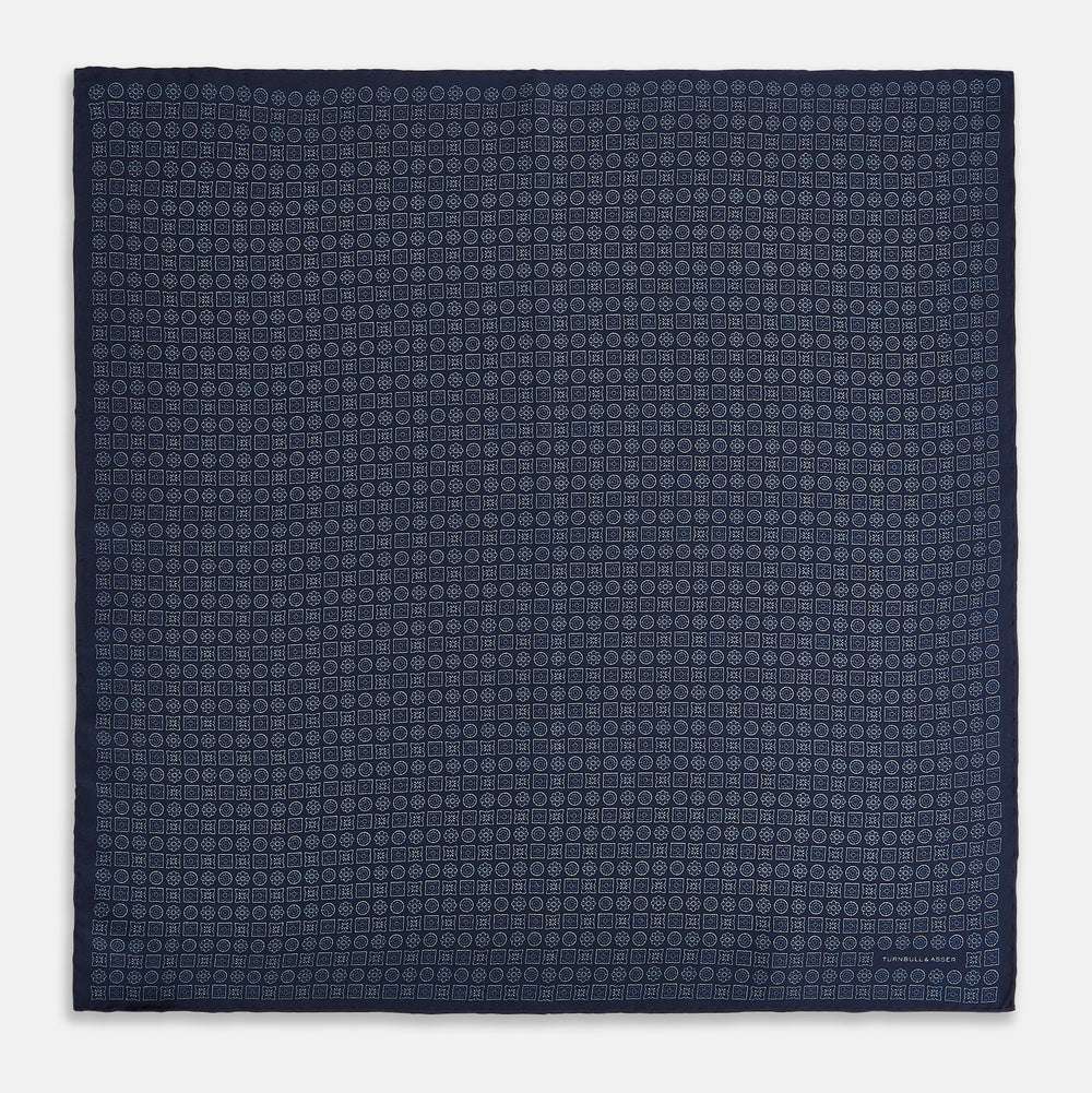A Floral Navy Pocket Square in Silk by Turnbull & Asser UK, featuring a repeating geometric pattern with the brand’s name printed in white in the corner—perfect for adding a touch of sophistication to formal tailoring. The top right edge is folded over to reveal the reverse side.