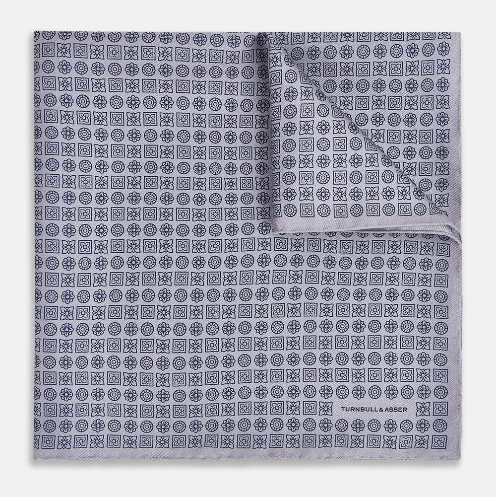 A lilac silk pocket square featuring a repeating geometric rosette pattern. One corner is folded over to show the reverse side, and "Turnbull & Asser UK" is printed in the bottom right corner.
