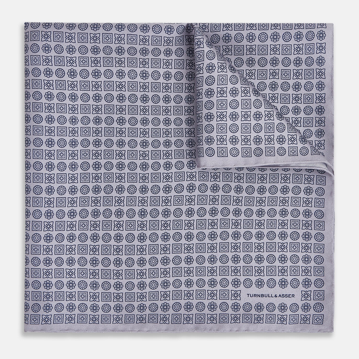 View 1 of product A lilac silk pocket square featuring a repeating geometric rosette pattern. One corner is folded over to show the reverse side, and "Turnbull & Asser UK" is printed in the bottom right corner.