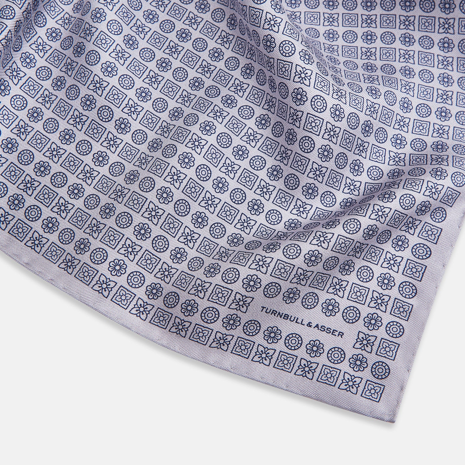 View 2 of product A lilac silk pocket square featuring a repeating geometric rosette pattern. One corner is folded over to show the reverse side, and "Turnbull & Asser UK" is printed in the bottom right corner.