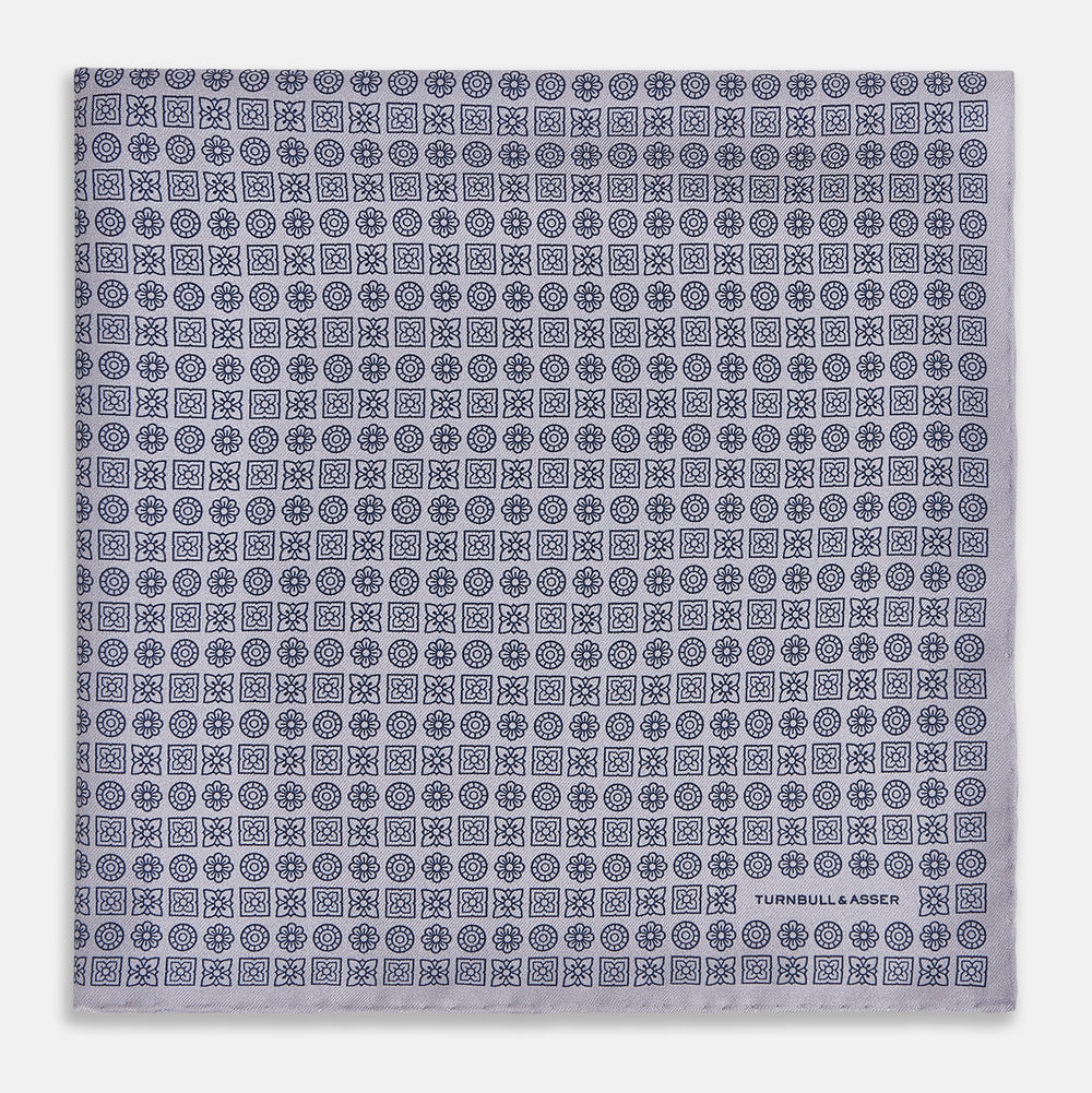 A lilac silk pocket square featuring a repeating geometric rosette pattern. One corner is folded over to show the reverse side, and "Turnbull & Asser UK" is printed in the bottom right corner.