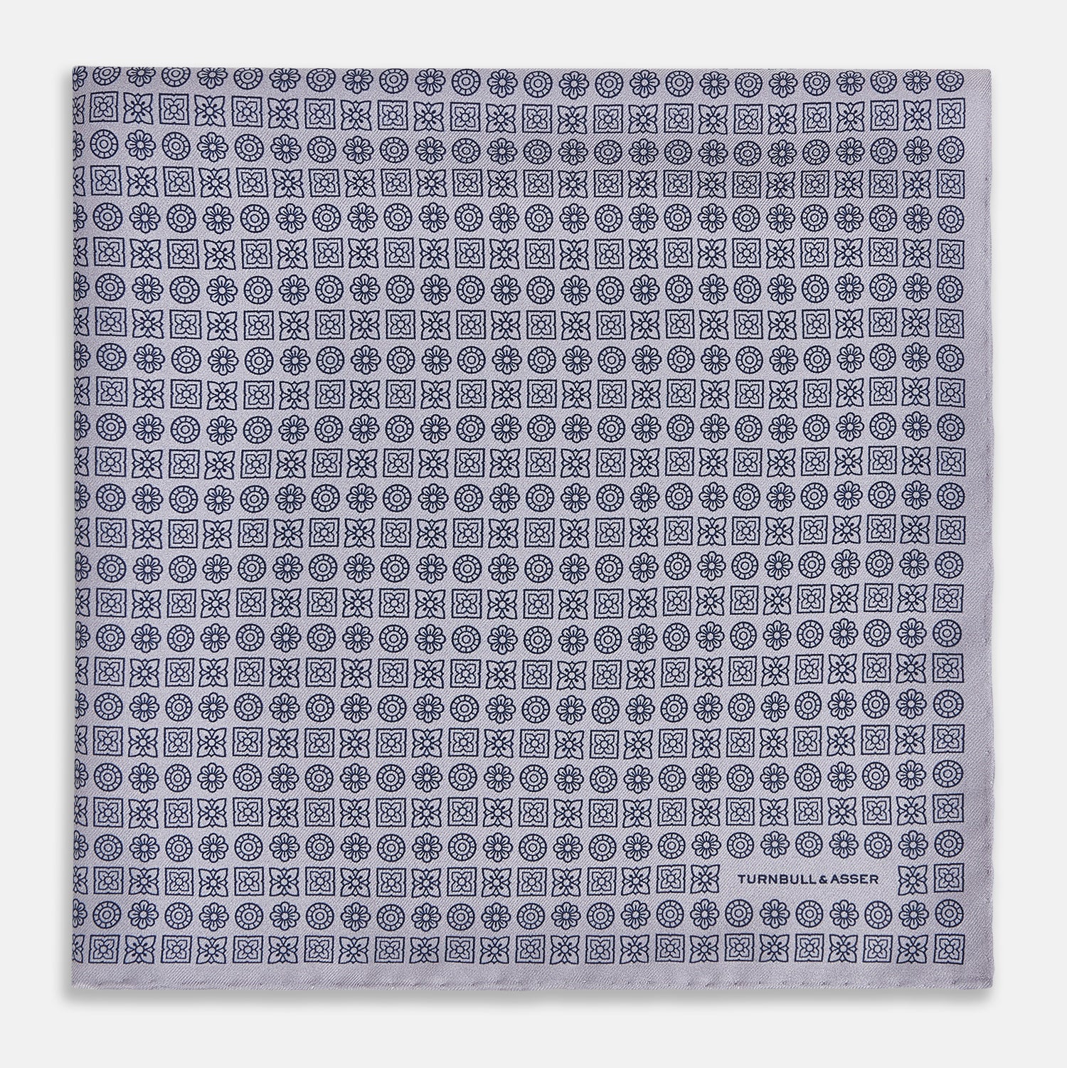 View 3 of product A lilac silk pocket square featuring a repeating geometric rosette pattern. One corner is folded over to show the reverse side, and "Turnbull & Asser UK" is printed in the bottom right corner.