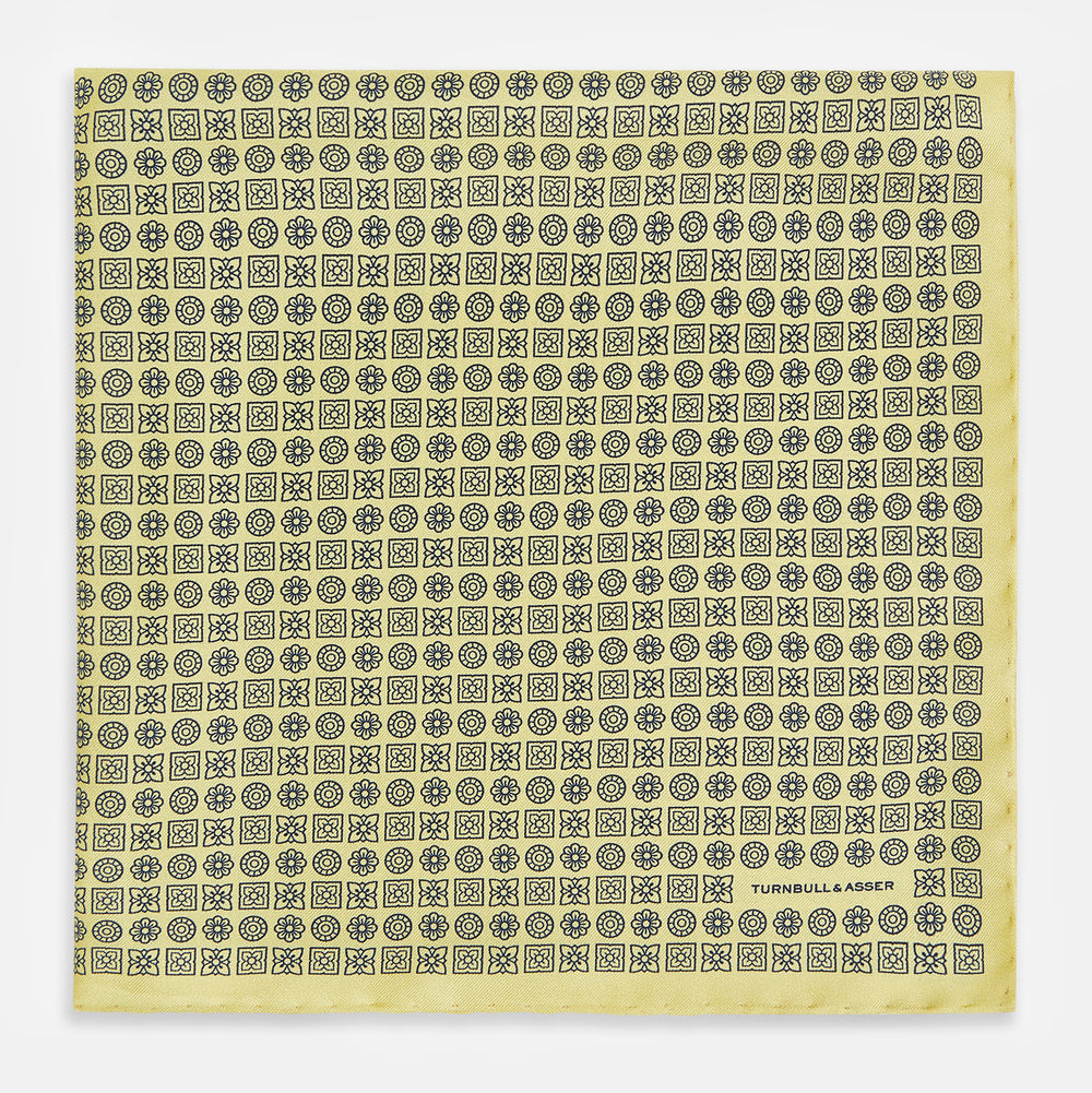 A Floral Yellow Pocket Square in Silk by Turnbull & Asser UK, showcasing a repeating black geometric pattern with subtle floral motifs and the brand logo in one corner. One corner of the fabric is folded over to display the reverse side.