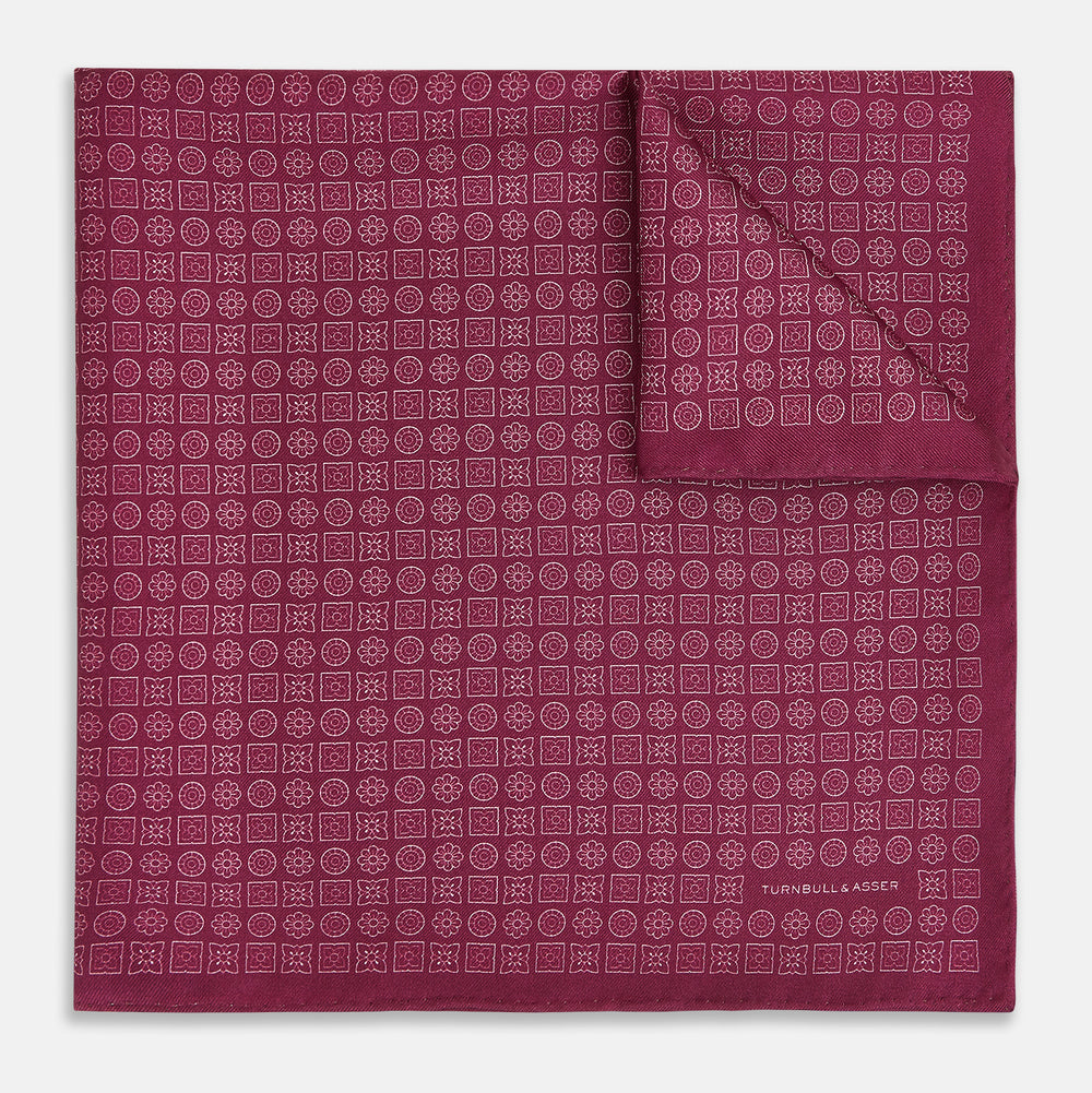 A folded Floral Burgundy Pocket Square in Silk by Turnbull & Asser UK, adorned with pink floral geometric motifs and featuring the Turnbull & Asser logo in the bottom right corner.