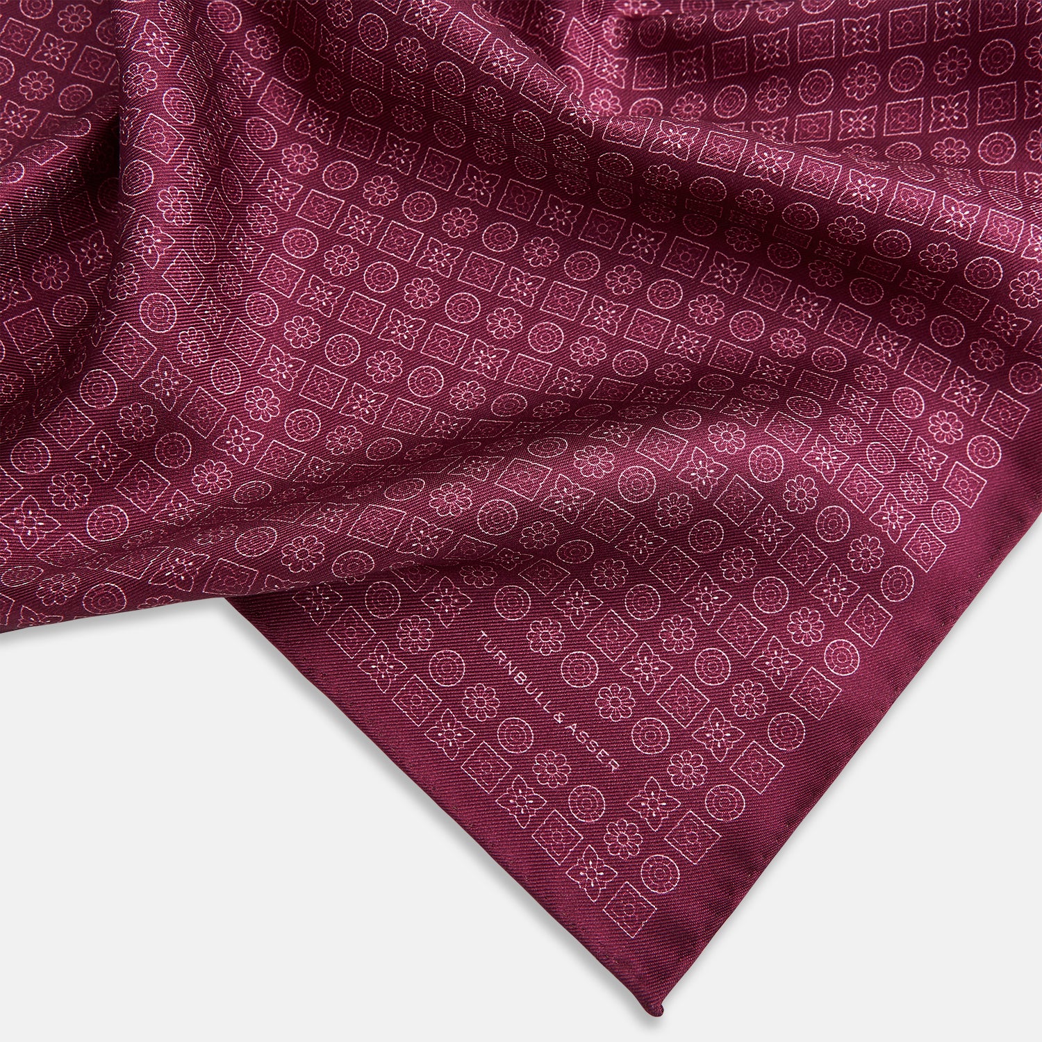 View 2 of product A folded Floral Burgundy Pocket Square in Silk by Turnbull & Asser UK, adorned with pink floral geometric motifs and featuring the Turnbull & Asser logo in the bottom right corner.