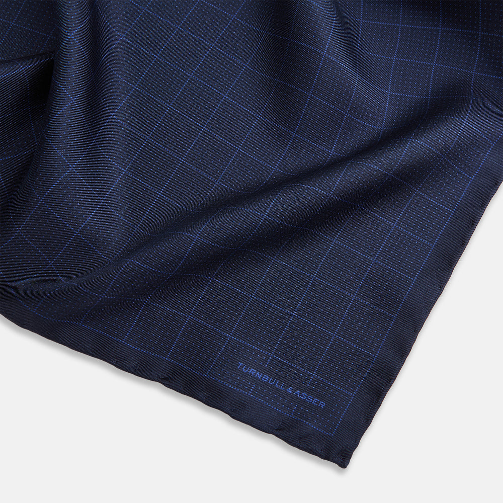 The Checked Blue On Navy Pocket Square in Silk by Turnbull & Asser UK is crafted from dark blue silk and features a subtle geometric design with a lighter blue grid pattern, finished with a discreet Turnbull & Asser logo in the corner.