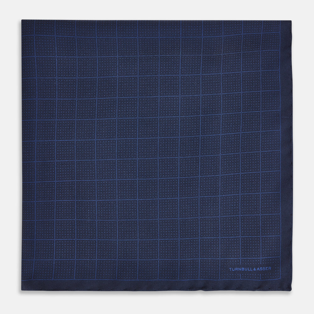 The Checked Blue On Navy Pocket Square in Silk by Turnbull & Asser UK is crafted from dark blue silk and features a subtle geometric design with a lighter blue grid pattern, finished with a discreet Turnbull & Asser logo in the corner.