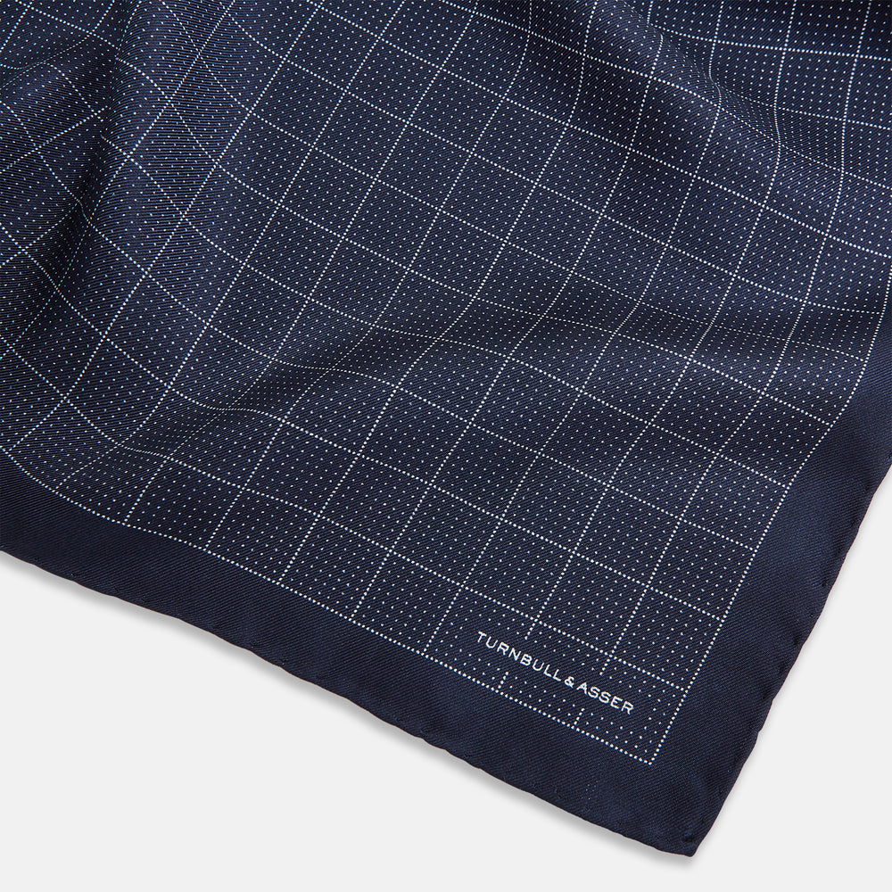 Checked Navy On White Pocket Square in Silk by Turnbull & Asser UK, featuring a navy blue geometric pattern with a white dotted grid and a solid border. The pocket square is shown with one corner folded over to display the “TURNBULL & ASSER” label in white text.