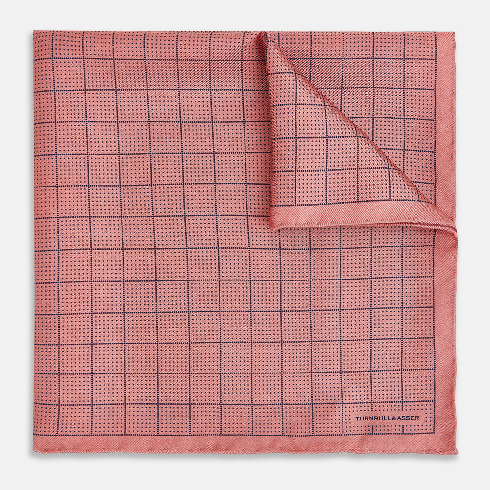 Introducing the Checked Pink Pocket Square in Silk from Turnbull & Asser UK, crafted with a geometric grid pattern of black lines and micro dots on soft pink silk. The design features a neatly folded corner and "Turnbull & Asser" subtly printed in the bottom right.
