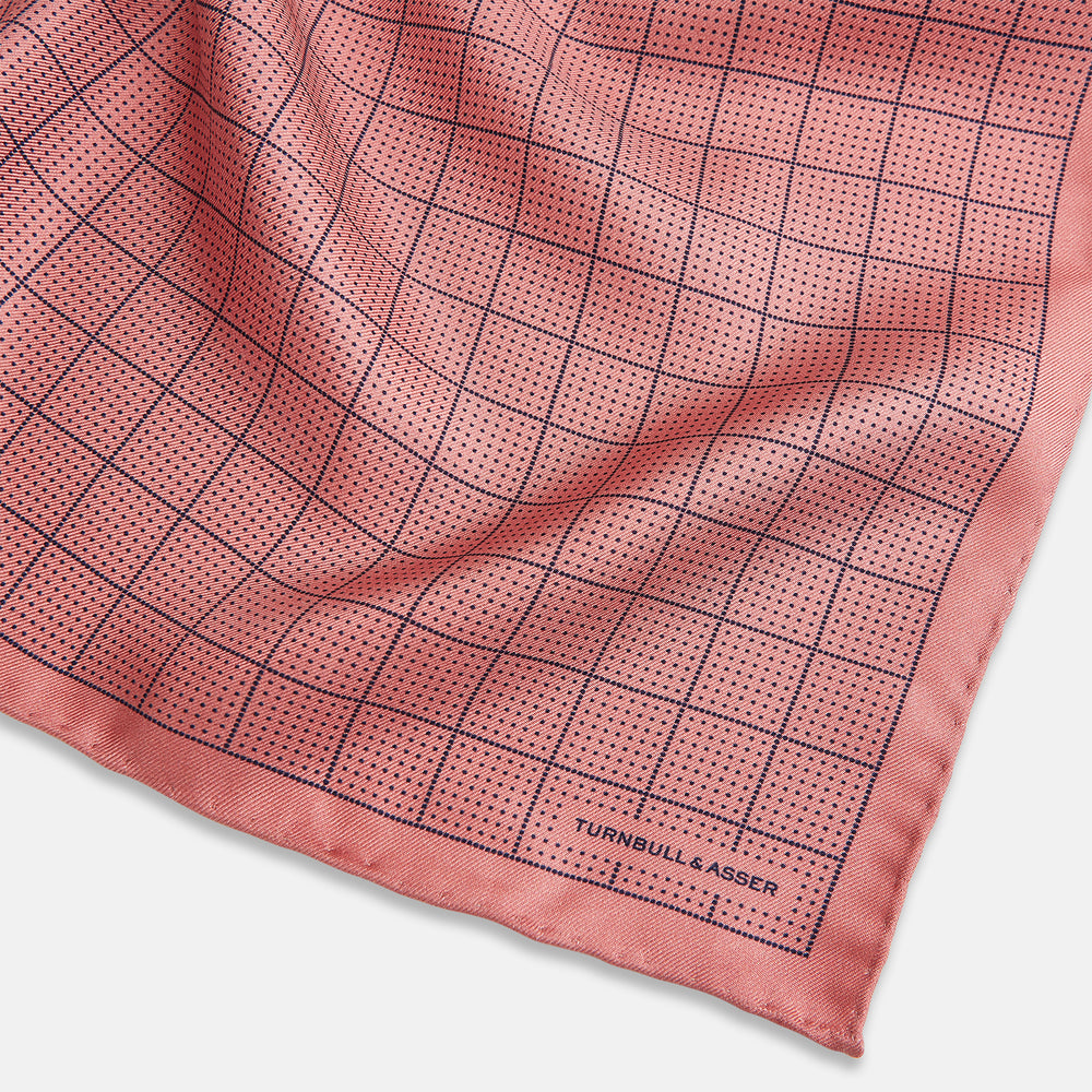 Introducing the Checked Pink Pocket Square in Silk from Turnbull & Asser UK, crafted with a geometric grid pattern of black lines and micro dots on soft pink silk. The design features a neatly folded corner and "Turnbull & Asser" subtly printed in the bottom right.