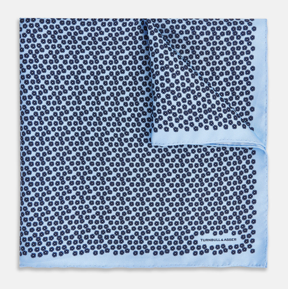 A light blue double-sided pocket square made of silk, featuring a dense pattern of small, dark blue floral motifs. One corner is folded over to reveal the Turnbull & Asser UK brand name printed in blue.