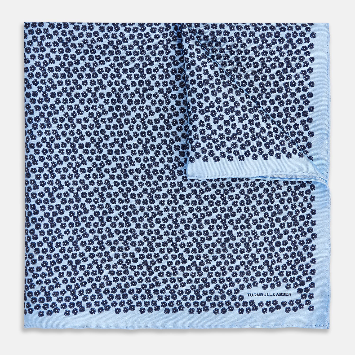 View 1 of product A light blue double-sided pocket square made of silk, featuring a dense pattern of small, dark blue floral motifs. One corner is folded over to reveal the Turnbull & Asser UK brand name printed in blue.