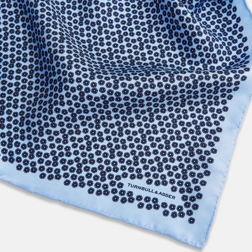 A light blue double-sided pocket square made of silk, featuring a dense pattern of small, dark blue floral motifs. One corner is folded over to reveal the Turnbull & Asser UK brand name printed in blue.