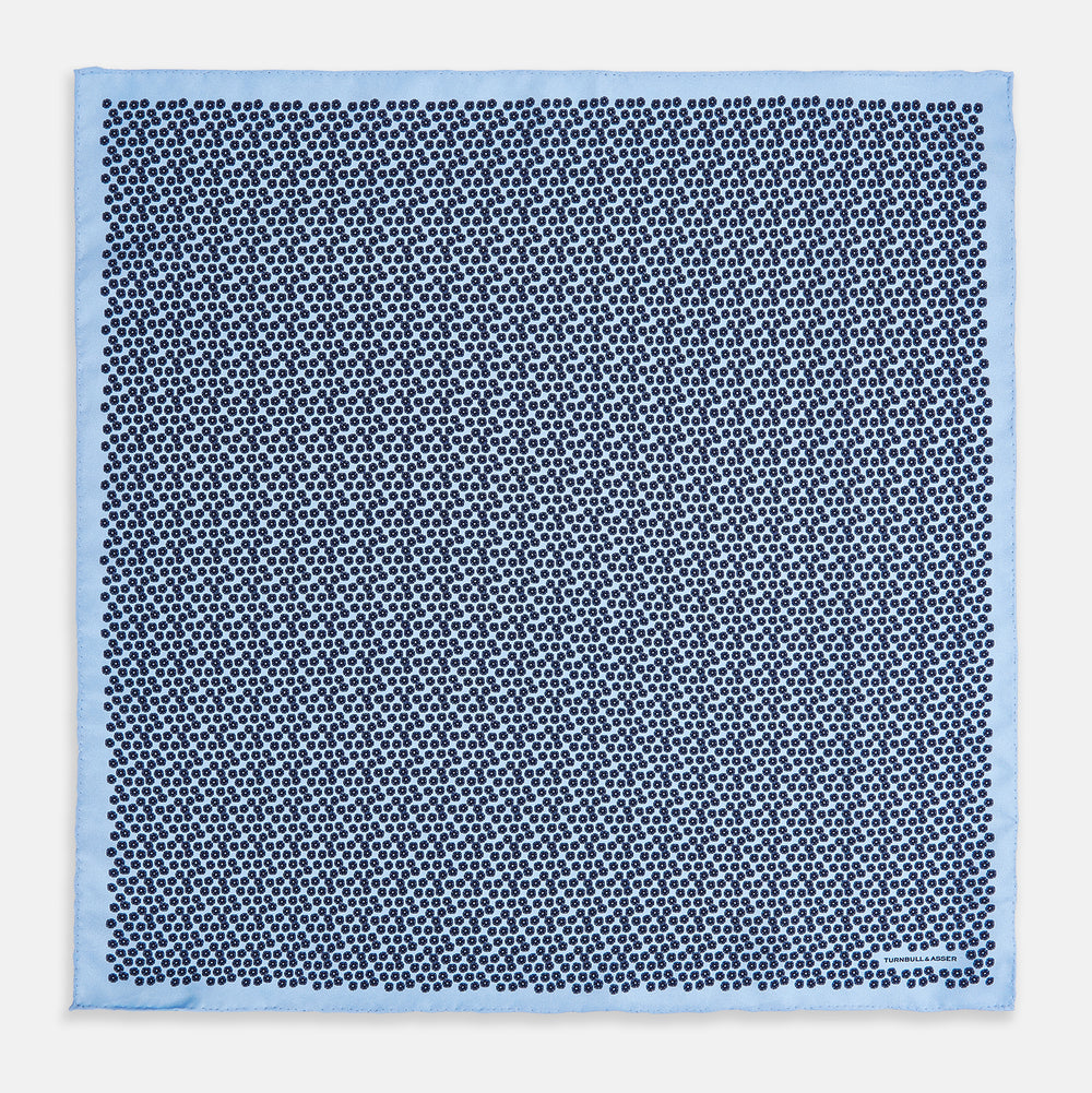 A light blue double-sided pocket square made of silk, featuring a dense pattern of small, dark blue floral motifs. One corner is folded over to reveal the Turnbull & Asser UK brand name printed in blue.