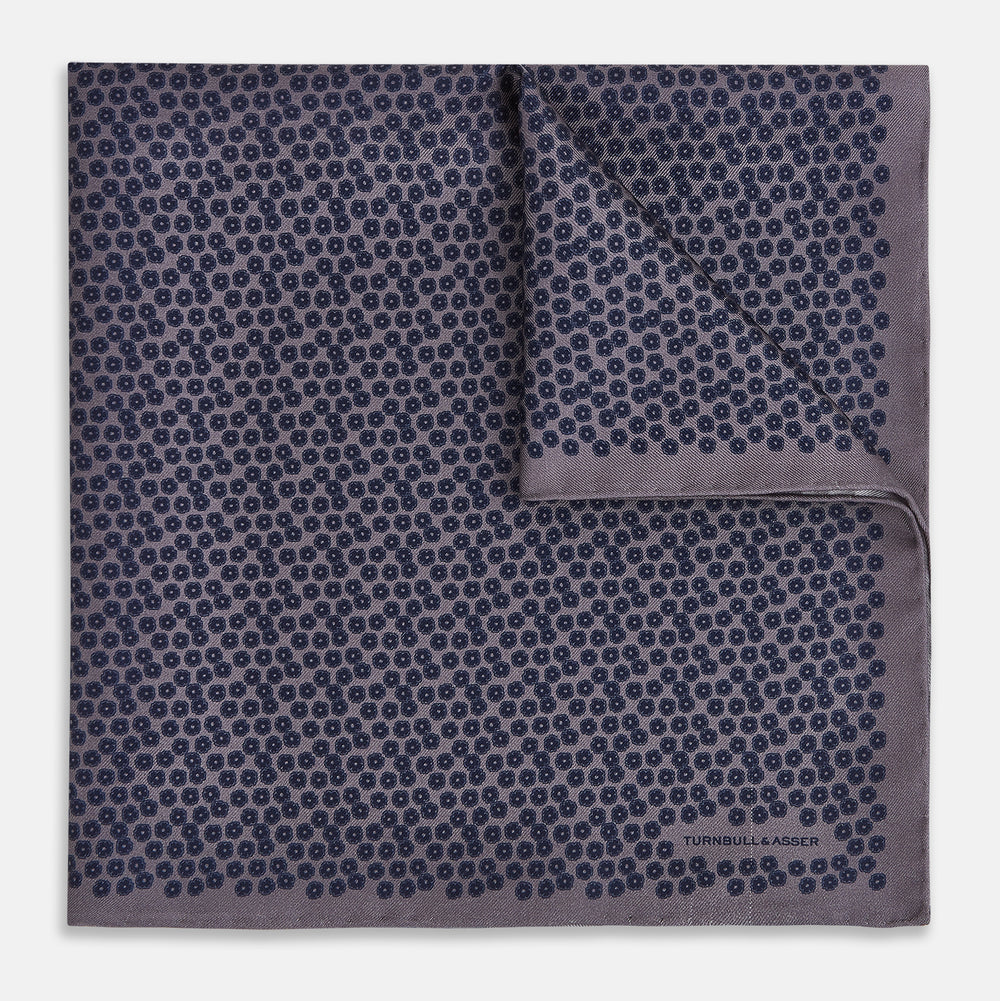 A folded Floral Purple Pocket Square in Silk by Turnbull & Asser UK, featuring a double-sided design with a pattern of small, dark blue circles and the brand’s logo printed in one corner.