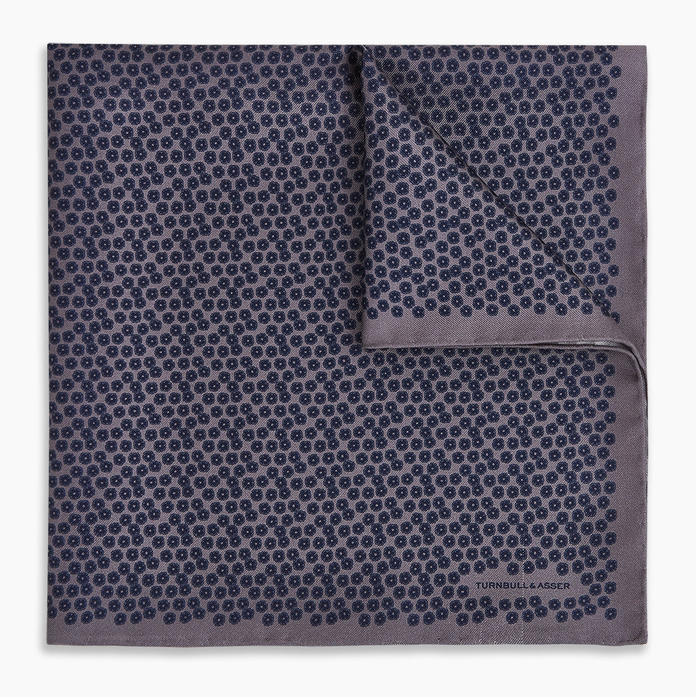 A folded Floral Purple Pocket Square in Silk by Turnbull & Asser UK, featuring a double-sided design with a pattern of small, dark blue circles and the brand’s logo printed in one corner.