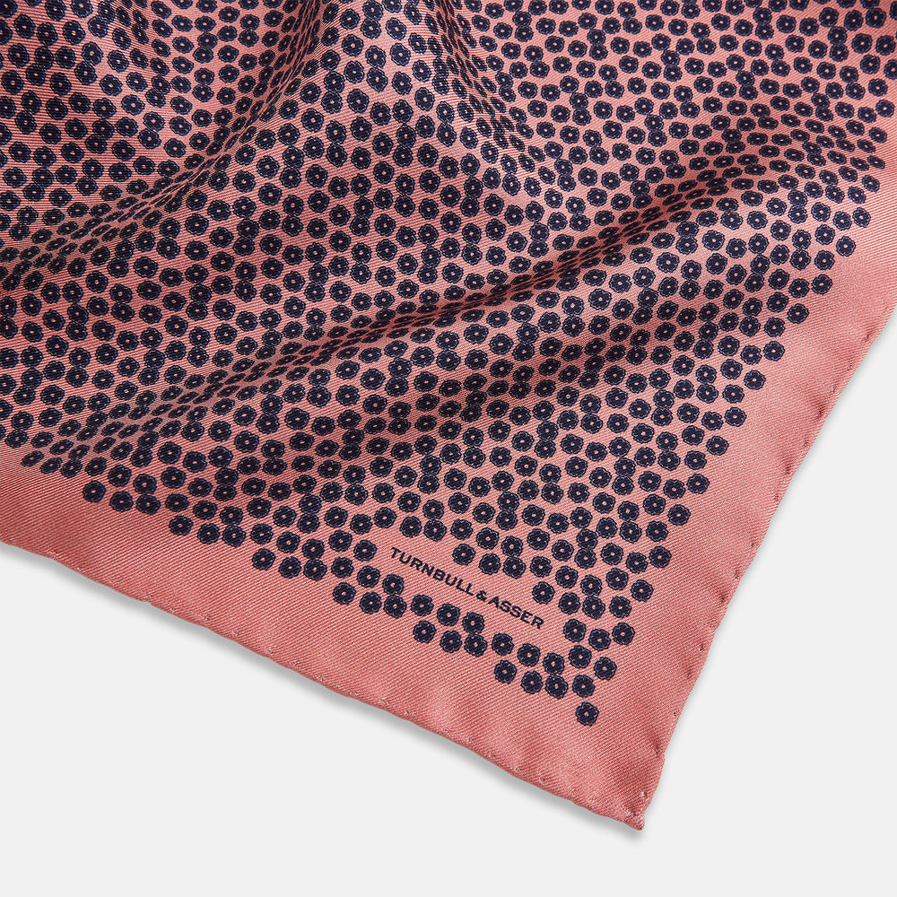 A folded Floral Pink Pocket Square in Silk by Turnbull & Asser UK, adorned with a pattern of small navy blue circles and the brand name printed in one corner, features a refined double-sided design for added versatility.