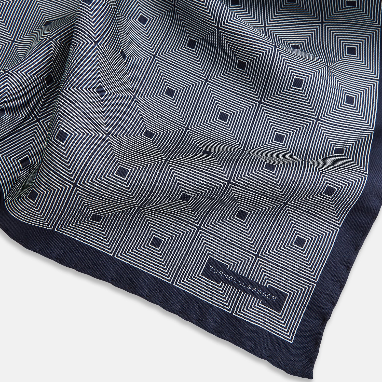 View 2 of product A Geometric Blue Pocket Square in Silk by Turnbull & Asser UK, featuring a navy blue and white geometric square tile design and a small label in the bottom right corner.
