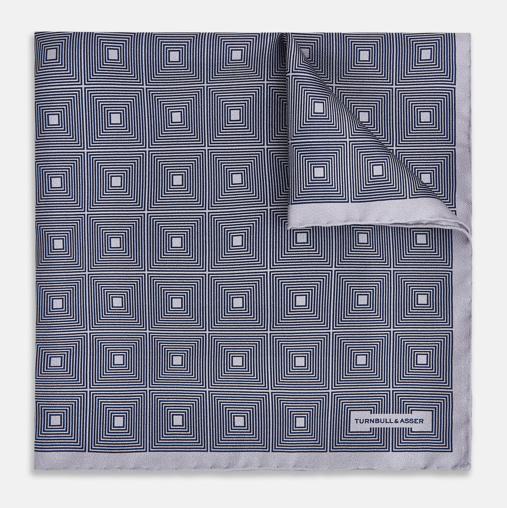 A folded, square Turnbull & Asser UK Geometric Grey Pocket Square in Silk featuring a geometric black and white concentric squares pattern accented with subtle navy lines, finished with a small Turnbull & Asser label in one corner.