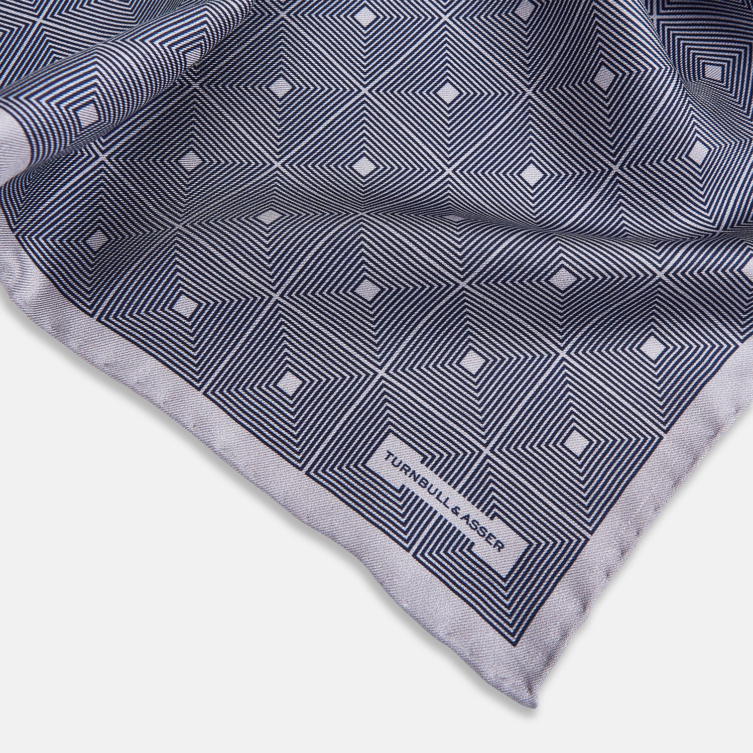 View 2 of product A folded, square Turnbull & Asser UK Geometric Grey Pocket Square in Silk featuring a geometric black and white concentric squares pattern accented with subtle navy lines, finished with a small Turnbull & Asser label in one corner.