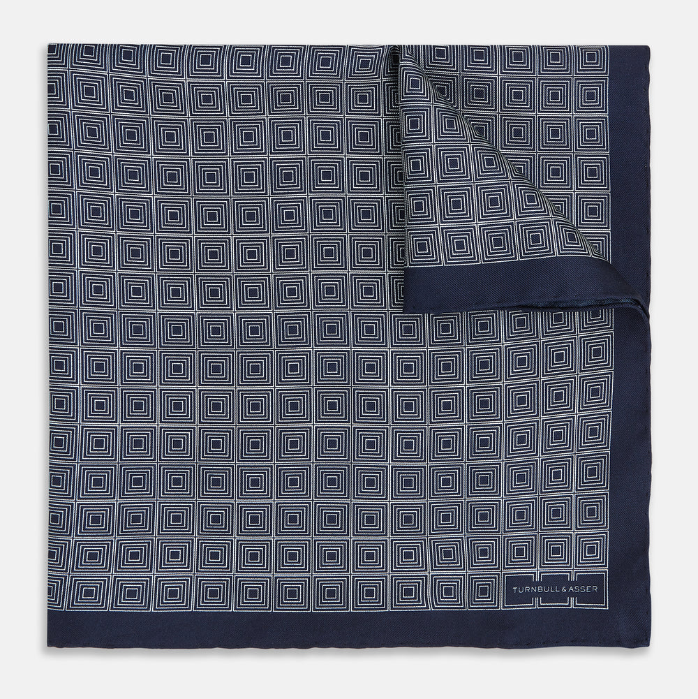 A Geometric Navy Pocket Square in Silk by Turnbull & Asser UK, showcasing a geometric pattern of small, overlapping concentric white squares and a stitched border. The pocket square is pictured with one corner folded over to display the Turnbull & Asser UK brand label.