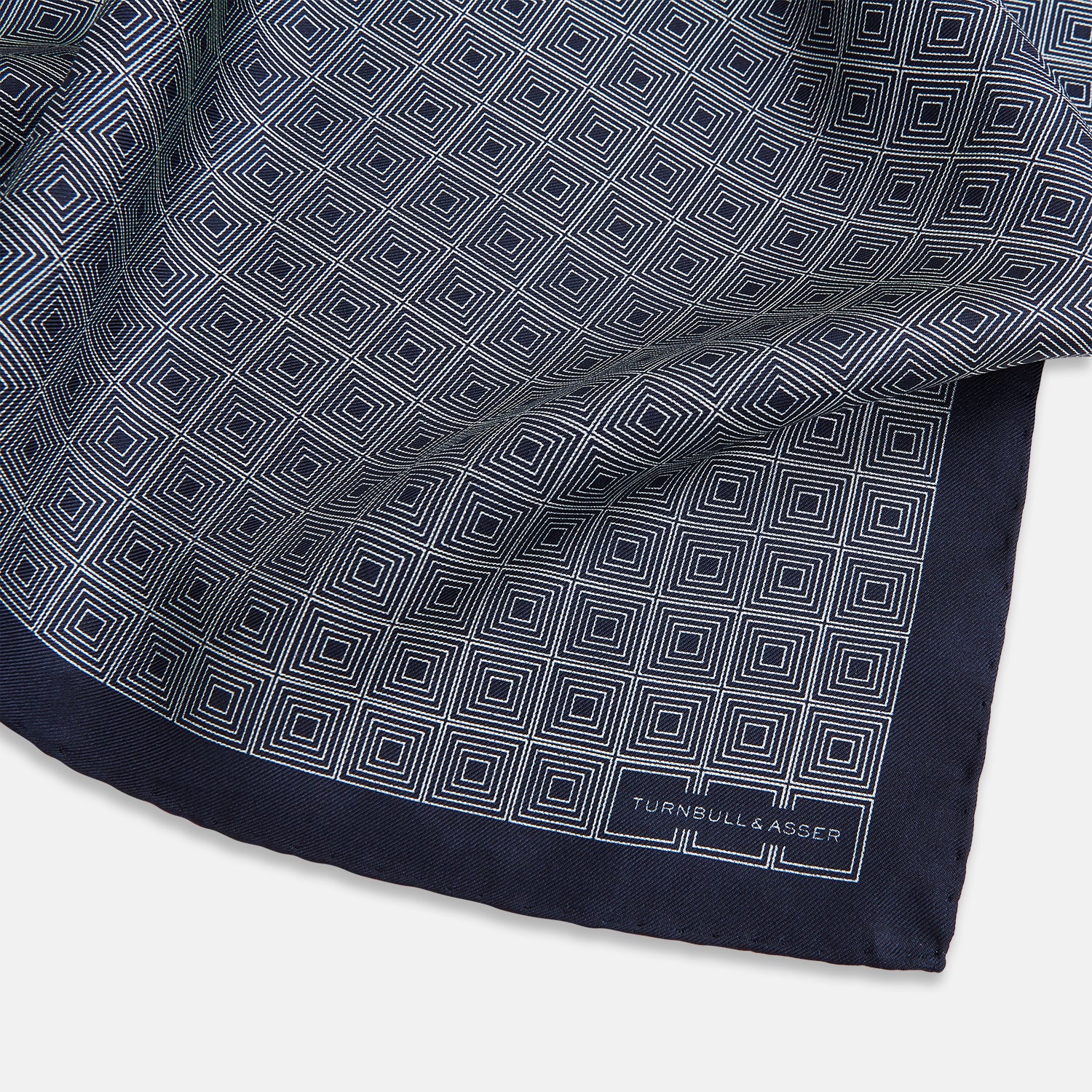 View 2 of product A Geometric Navy Pocket Square in Silk by Turnbull & Asser UK, showcasing a geometric pattern of small, overlapping concentric white squares and a stitched border. The pocket square is pictured with one corner folded over to display the Turnbull & Asser UK brand label.