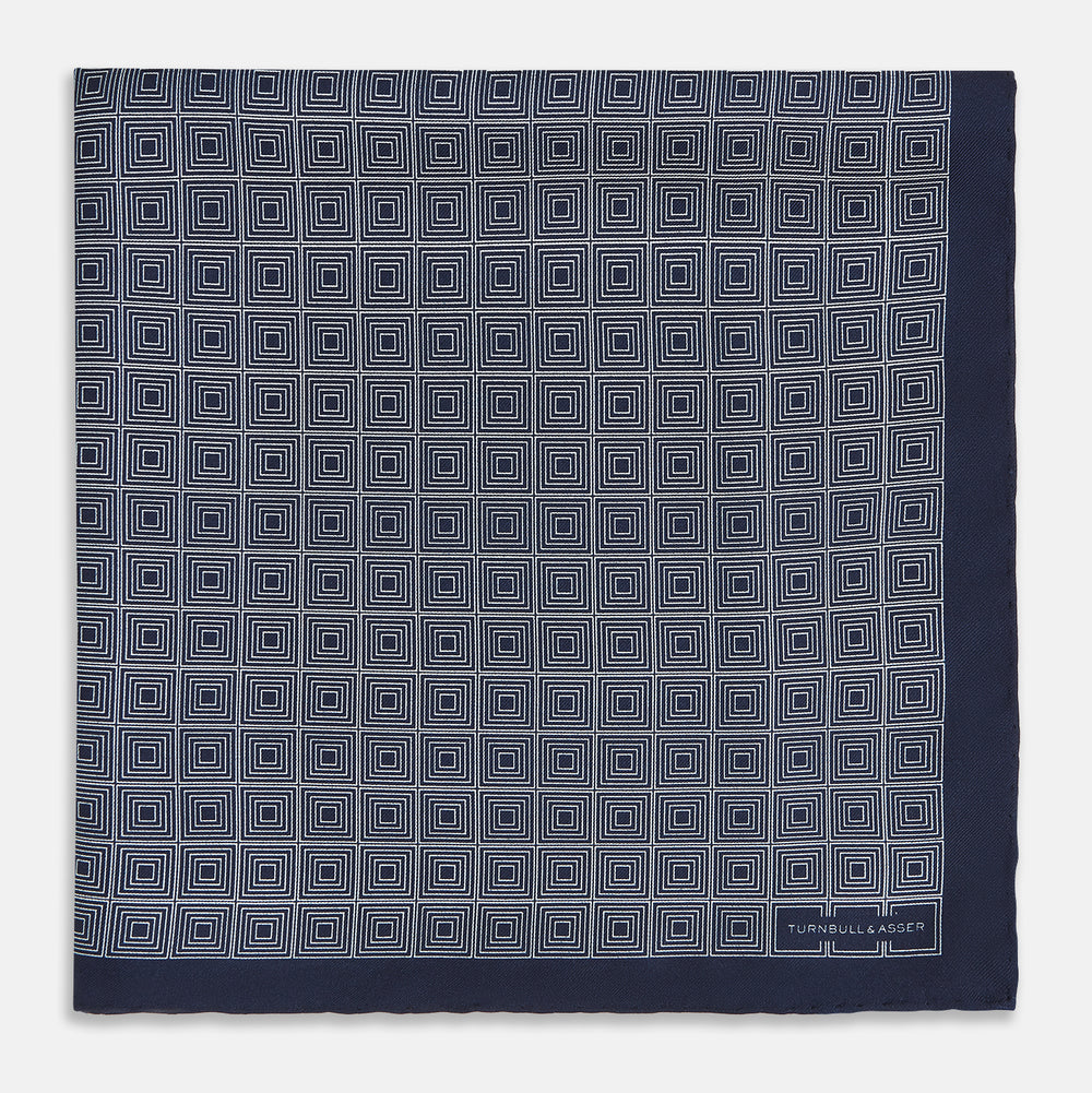 A Geometric Navy Pocket Square in Silk by Turnbull & Asser UK, showcasing a geometric pattern of small, overlapping concentric white squares and a stitched border. The pocket square is pictured with one corner folded over to display the Turnbull & Asser UK brand label.