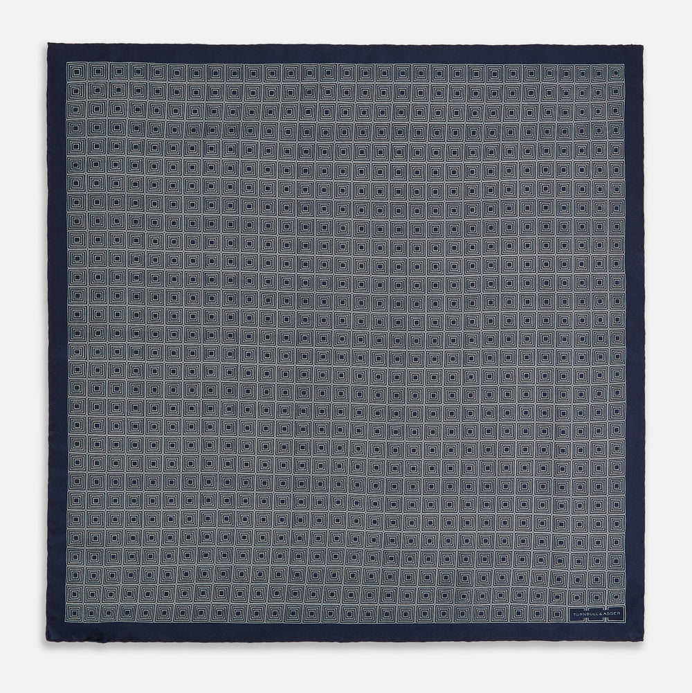 A Geometric Navy Pocket Square in Silk by Turnbull & Asser UK, showcasing a geometric pattern of small, overlapping concentric white squares and a stitched border. The pocket square is pictured with one corner folded over to display the Turnbull & Asser UK brand label.