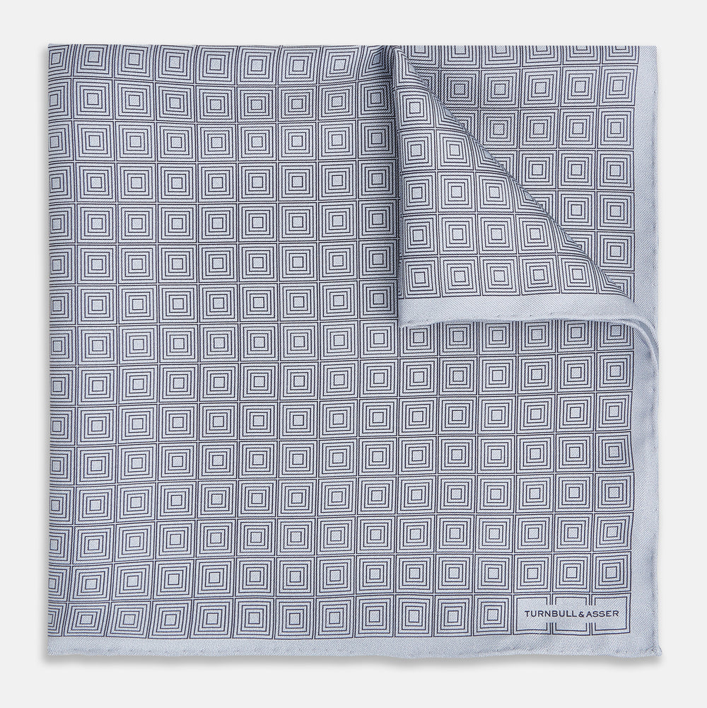 Geometric silver pocket square in silk by Turnbull & Asser UK, featuring a striking pattern of dark blue concentric squares. One corner of the fabric is folded to reveal the reverse side, with the brand name Turnbull & Asser UK printed elegantly in one corner.