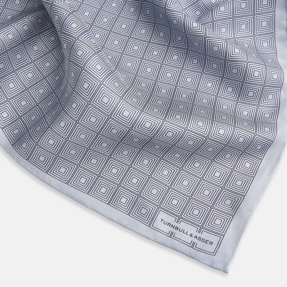 Geometric silver pocket square in silk by Turnbull & Asser UK, featuring a striking pattern of dark blue concentric squares. One corner of the fabric is folded to reveal the reverse side, with the brand name Turnbull & Asser UK printed elegantly in one corner.
