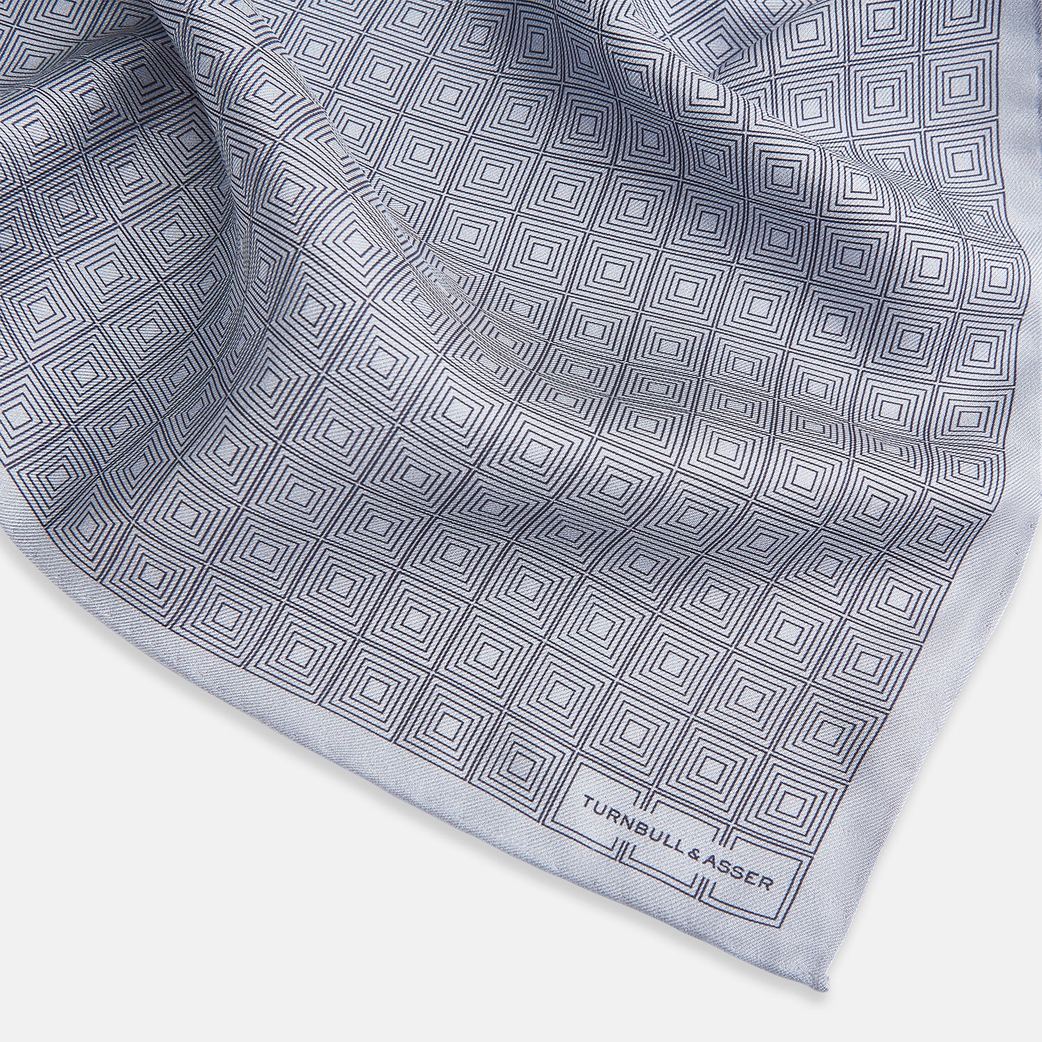 View 2 of product Geometric silver pocket square in silk by Turnbull & Asser UK, featuring a striking pattern of dark blue concentric squares. One corner of the fabric is folded to reveal the reverse side, with the brand name Turnbull & Asser UK printed elegantly in one corner.