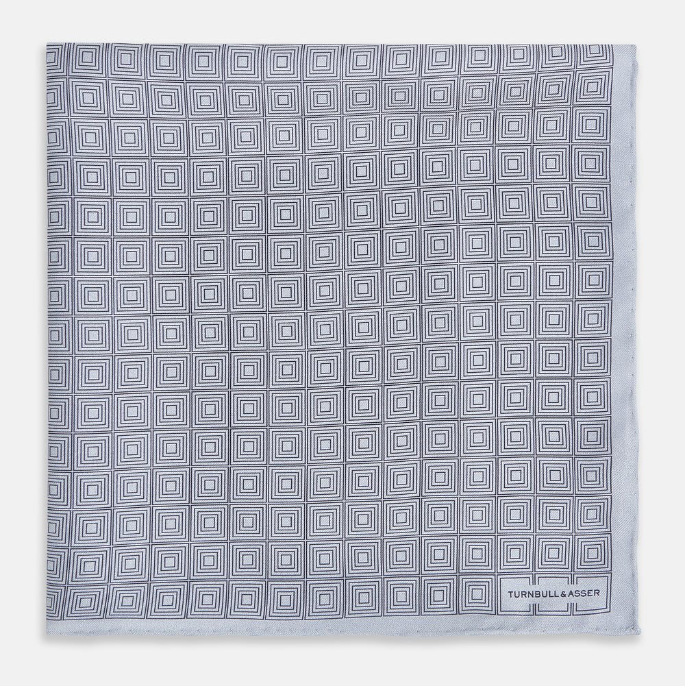 Geometric silver pocket square in silk by Turnbull & Asser UK, featuring a striking pattern of dark blue concentric squares. One corner of the fabric is folded to reveal the reverse side, with the brand name Turnbull & Asser UK printed elegantly in one corner.