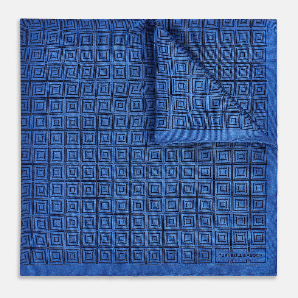 The Geometric Blue Pocket Square in Silk by Turnbull & Asser UK features a vibrant blue design adorned with small, outlined squares arranged in a grid of sharp navy lines. One corner is neatly folded to reveal the reverse side, with "Turnbull & Asser" printed at the bottom right.